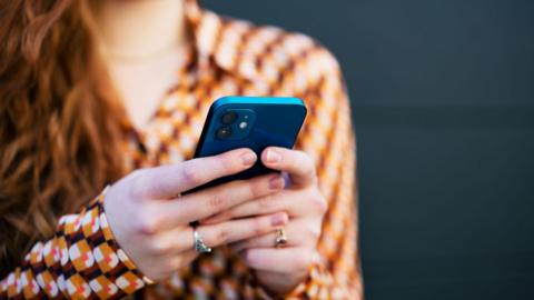 A smartphone being read, held in the hands of a woman with long, wavy, red hair and wearing a brown, orange, white and red-patterned shirt.