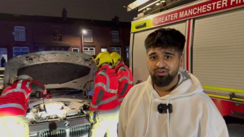 Nizam Pate stands in front of his grey BMW. The bonnet is up and the firefighters are stood over it while. Nizam has short dark hair, he wears a white hoodie. A fire engine can be seen in the background.