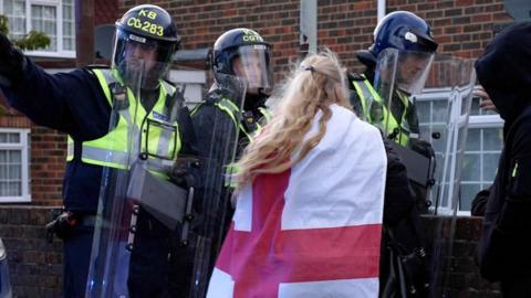 A woman with an England flag around draped around her is surrounded by policemen holding shields 