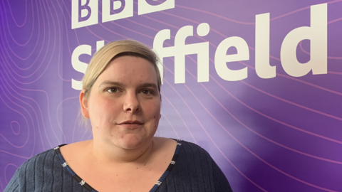 A woman with blonde, tied-back hair stands in front of a BBC Sheffield logo board.