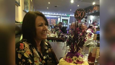 Maria Britton on her 50 birthday, in a restaurant with a 50 balloon and a birthday cake. She is smiling and wearing a floral top.