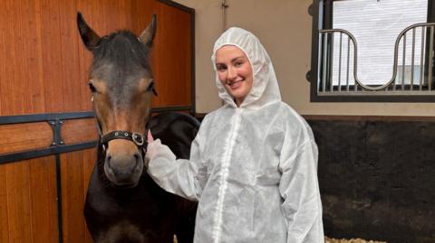 A woman wearing a zipped up white boiler suit, with the hood covering her hair, stands next to a chestnut brown pony, wearing a bridle over the bridge of its nose. They are in a stable with hay on the floor.