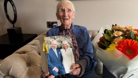 An elderly woman with white hair, wearing a blue cardigan and a patterned scarf, sits on a cream sofa holding a large card featuring a photograph of King Charles and Queen Camilla. A bouquet of orange, yellow and peach flowers sits beside her.