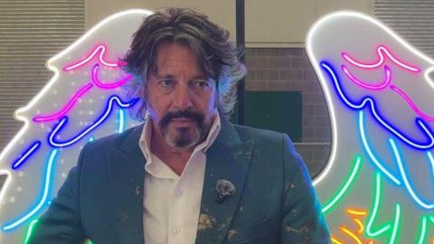 Laurence Llewelyn-Bowen stands in front of neon angel wings. He has long dark hair and a black and grey beard. He wears a white shirt and a green blazer