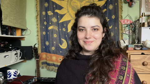 Lily poses for a photograph next to her recording and mixing equipment in her bedroom in Leominster. Behind her are shelves of electronic equipment, a tapestry with the Sun, moons and stars, and a guitar.