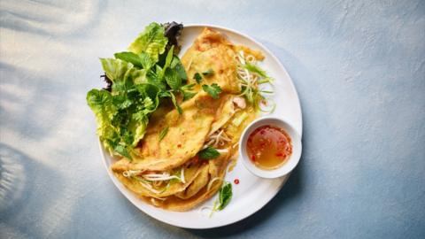 Top down view of banh xeo (Vietnamese panckaes) with some salad and a chilli dip on a white plate