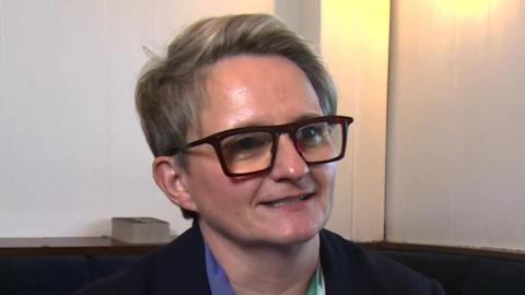 Charlotte Cadden speaks during a TV interview inside a room. She has short fair hair and wears dark glasses.