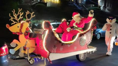 Father Christmas waving from a sleigh pulled by a wooden reindeer whilst a snowman carrying a charity bucket walks behind