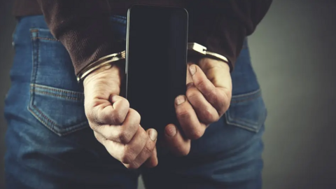 A person in a black top and jeans holds a phone behind their back while their hands are handcuffed.