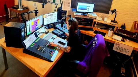A women sits on a large chair at a desk with radio broadcasting equipment in front of her. She is looking at two screens and touching buttons on the mixing desk. There are microphones, a record player, speakers and, lights and other equipment around the desk.