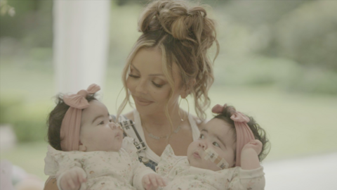 A woman with brown/blonde hair piled in a bun on top of her head. she is holding one brunette baby in each arm and gazing down at one of them. They are all wearing white and the babies - twin girls - both have a feeding tube in their nose and are wearing a pink bow on their heads.