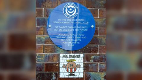 the wall where there is a blue plaque which shows the Portsmouth Football Club crest above text which reads "on this site once more stands a mighty football club. We cannot change the past but we can shape the future. Dedicated to all those fans who took a stand and refused to allow Portsmouth FC to die" underneath the plaque is a white mosaic with a mr man character dressed in a southampton fc kit, it says "mr saints" at the top