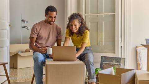 Smiling woman using laptop sitting by man with coffee cup in new apartment. Mid adult couple is planning together in new house. They are moving home.