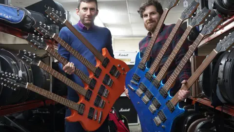 A blurred image of two men standing next to each other in a music shop, holding blue and red five-neck guitars.