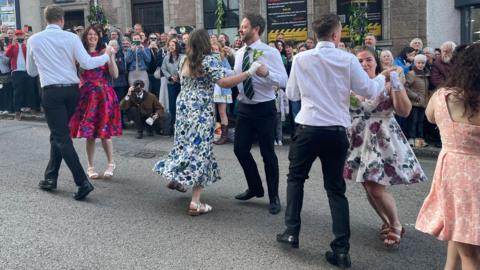 Four couples are dancing in the street. The pavements are lined with spectators.