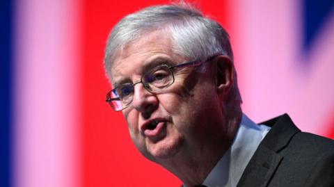 Mark Drakeford speaking while looking off camera to the left of the frame. He is swearing a suit and shirt, and glasses.