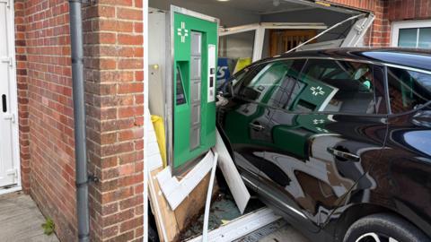 A black car has crashed into a red brick building. The door is caved in.