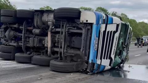 A blue and white lorry lying on it's side on the motorway carriagewat with it's wheels on show and it's windscreen smashed. a pool of diesel can be seen on the ground.