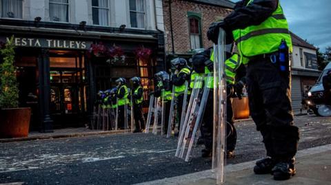 Police officers in riot gear stand in a line in the middle of a road in Sunderland. A pub is on the left, with a sign saying 'Vesta Tilleys'.