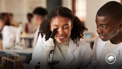A picture of a female looking into a microscope with a male student next to her, both wearing white lab coats.