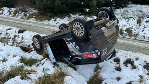 Hollie McRae's Mini lying upside down in a small ditch, surrounded by snow and ice.