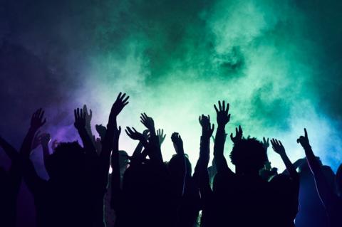 Silhouette of a crowd of people with their hands up, against green and purple lights and smoke.