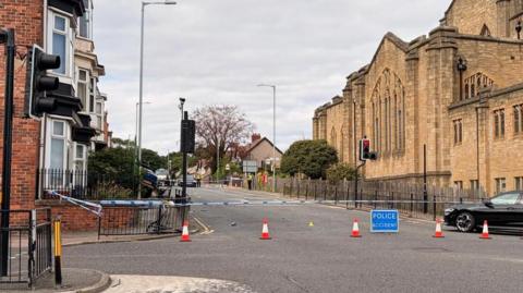 A police cordon near the junction between Kayll Road and Chester Road in Sunderland. The cordon, which is enforced with police tape and a blue police sign, shuts off a street with homes on the left side and a church on the right. To the left, part of a car can be seen in the bushes of a house. The railings near it are bent, implying it has smashed through them and into the house.