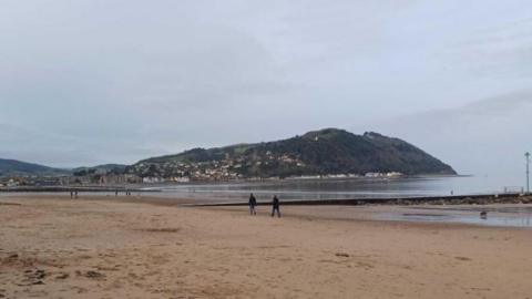 Minehead beach with some people walking and calm seas. North Hill is in the background.