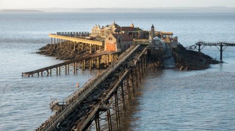 Birnbeck Pier in Weston-super-Mare. The pier is damaged and in disrepair, with a heavily damaged wooden walkway and dilapidated buildings. 