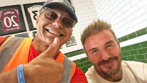 Pesh Kapashiawala is wearing an orange hi-vis vest over a red T-shirt. He is smiling and giving a thumbs up next to Sir David Beckham, who is wearing a white T-shirt and sitting down inside the restaurant. Sir David is also smiling.