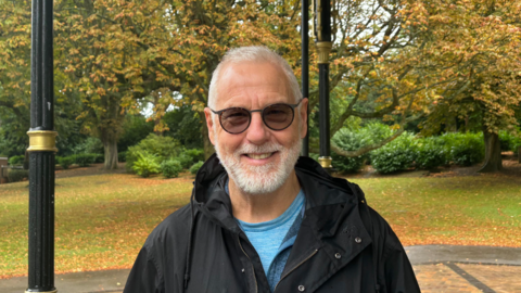 Campaigner smiles at the bandstand in Cannon Hill Park. He is wearing a blue top and black jacket. 