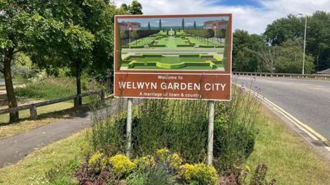 A brown sign showing a picture of a formal garden above the words "Welcome to Welwyn Garden City, a marriage of town and country" in white letters. It is set in a bed of shrubs surrounded by grass with a tree-lined path on one side and a road with double yellow lines on the other.
