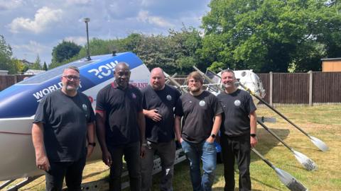Frank Bruno stands with the group that are rowing the Atlantic, with the boat behind them and oars to one side. Left to right, the men seen in the picture are Rob Oliver, Matt Humphries, Tom Hayward and Kev Joynes.