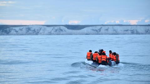 People in orange life-jackets on a small boat heading towards white cliffs that are in the background