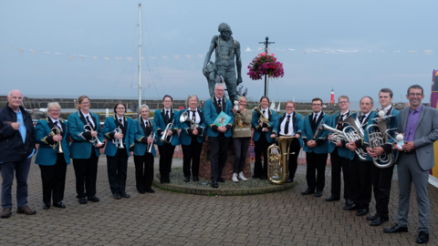 The band lined up on Watchet Harbour wearing blue jackets, black trousers and standing with their instruments. 