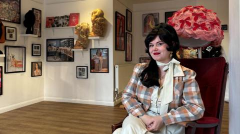 Sarah Wood on the right - she is sitting down, has black hair, red lipstick, is wearing a cream dress and scarf and checked jacket with her exhibition including wigs and framed photographs behind her. 