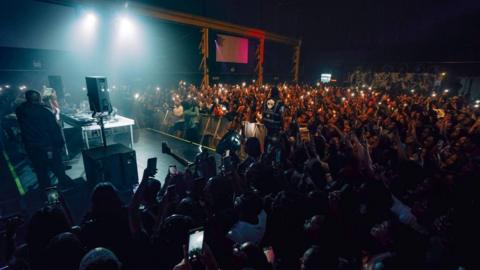 A huge crowd of people inside a music venue, some holding their phones aloft, watching a DJ at their decks, with barriers separating them from the crowd. Speakers can be seen either side of the decks.