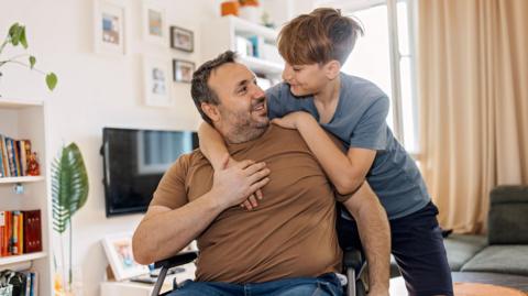 A boy gives his father, who is a wheelchair user, a hug in their home