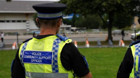 The back of a police community support officer who is stood on a field. He is wearing a black hat, black t-shirt and a high visibility vest with his job title on