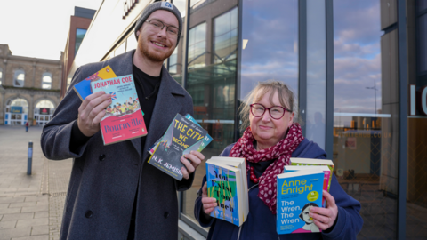 City of Wolverhampton Council cabinet member for city development, jobs and skills, councillor Chris Burden and Gently Used Book Club owner, Pat Austin, outside the Ignite business hub. Both are holding books in their hands and smiling at the camera. Chris has a long grey coat on and hat. Pat has a patterned scarf on over a blue coat.