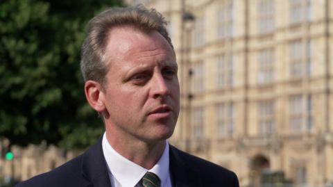 Joe Robertson MP gives a TV interview outside the Houses of Parliament. He has light brown, swept back hair and wears a dark suit, white shirt and tie.