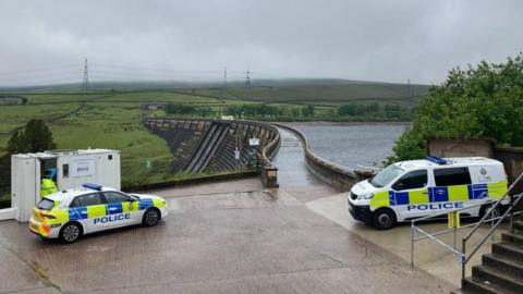 Police vehicles in the foreground, with a dam and reservoir in the background.