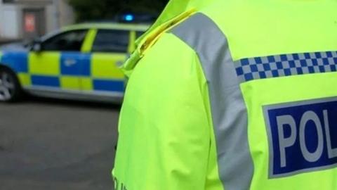 A police officer wearing a yellow hi-vis jacket with his back to the camera and a police car with yellow and blue markings in the background