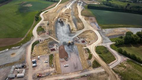 An aerial picture shows a major road construction site in the middle of green fields. A huge amount of groundwork has been undertaken, with a number of dirt roads encircling the site. Work vehicles, including white vans and yellow trucks, appear tiny.