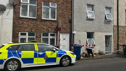 A bright yellow and blue police car with a star logo is parked next to a grey terrace house which has a white and blue striped police cordon draped across it. All the doors and windows of the property are open.