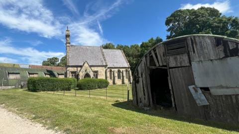 A small stone church with green hedges to the front stands among farm buildings, some in a dilapidated condition, on a bright sunny day.