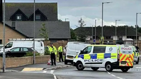 A police van parked across a road in Dumfries. The van is white with bright green and blue markings down the side and the word police in blue. Two officers in hi viz jackets are on the pavement to the left.