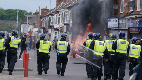 A line of police officers facing a burning car.