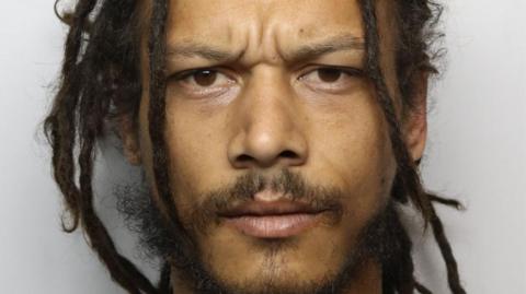 A mugshot image of a man with dark brown dreadlocks, a moustache and goatee frowning at the camera