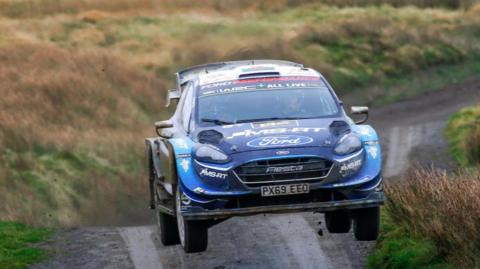 A blue world rally car with all four wheels off the ground as it moves at speed over a crest on a rally stage
